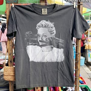 May include: Dark gray t-shirt featuring a black and white graphic of a woman's face with hands covering her mouth. The shirt has the text "Pepper Comfort Colors" at the top and "Project X Creative" at the bottom.