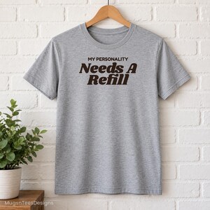 May include: A heather grey short-sleeved t-shirt featuring the dark brown text MY PERSONALITY Needs A Refill printed on the chest. The shirt has a crew neckline and is displayed on a wooden hanger against a white brick wall background.