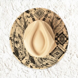 May include: Tan fedora hat with a decorative brim featuring black feather and geometric patterns. The hat has a beige crown and a wide brim, perfect for sun protection and fashion.