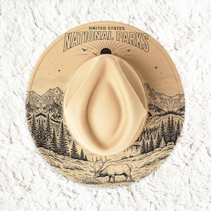 Puede incluir: Un sombrero fedora de color beige con una ilustración en tinta negra de un paisaje montañoso, pinos y un alce. El texto UNITED STATES NATIONAL PARKS está impreso en negro en la parte superior, junto a un motivo de sol y pequeñas estrellas.