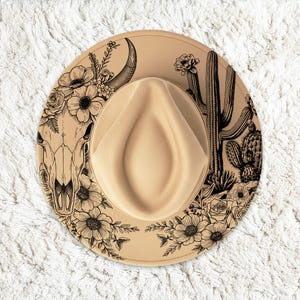 Puede incluir: Sombrero fedora color beige con un ala decorativa con un diseño floral y desértico en blanco y negro. El diseño incluye una calavera de toro, cactus y flores. El sombrero tiene una corona clásica en forma de lágrima.