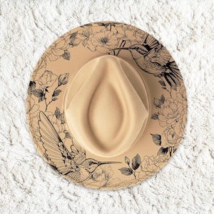 May include: A tan fedora hat with a cream-coloured crown. The brim features a black floral and hummingbird design. The hat is set against a white, textured background.