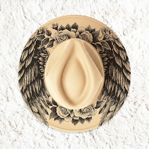 May include: A beige fedora hat with a decorative design. The hat features black angel wings and floral rose patterns around the brim. The hat is a neutral color and is suitable for various outfits.
