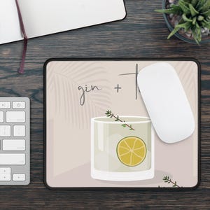 May include: A rectangular mousepad featuring a cocktail illustration. The design includes a glass with a lemon slice, sprigs of herbs, and the word "gin." A white computer mouse rests on the pad.