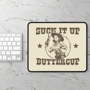Puede incluir: Alfombrilla de ratón rectangular con fondo beige y borde negro. El diseño presenta una ilustración vintage de una mujer sosteniendo un arma, con el texto "SUCK IT UP BUTTERCUP" en marrón, junto con dos estrellas.