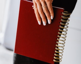 Könnte beinhalten: Ein rotes Spiralnotizbuch mit goldenen Ringen. Das Notizbuch ist geschlossen und wird von einer Hand mit goldenen Ringen gehalten.