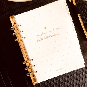 May include: A white planner with gold accents and a gold heart. The planner has a dotted pattern and the text "new plans, new dreams, NEW BEGINNINGS" in gold lettering. The planner also has a gold tab for each month of the year. The planner is closed and lying on a black surface.