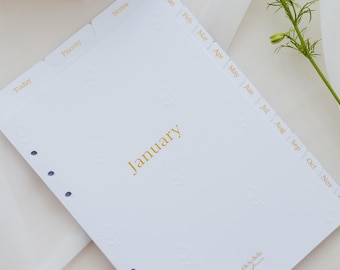 Op de afbeelding: Een witte planner-inlegvel met gouden tekst January, voorzien van maandelijkse tabbladen van Jan tot Nov en extra tabbladen voor Today, Priority en Notes. Het papier heeft een subtiel reliëf van bladeren en zes ronde perforatiegaten.