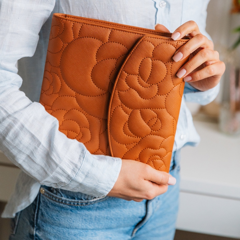 Girl is holding in her hand brown vegan leather portfolio, she is wearing blue jeans and blue shirt. Portfolio have floral designs and gold button.