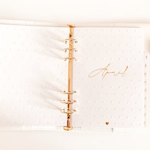Op de afbeelding: Een witte planner met gouden ringen en een gouden inscriptie met de tekst "April". De planner heeft een witte achtergrond met kleine witte stippen.