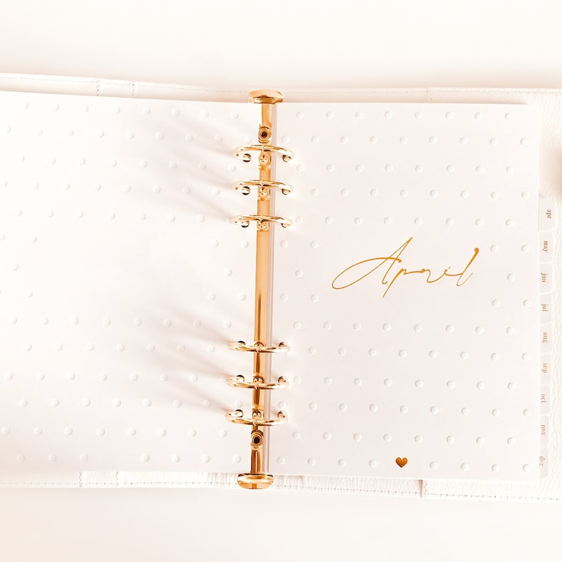 Op de afbeelding: Een witte planner met gouden ringen en een gouden inscriptie met de tekst "April". De planner heeft een witte achtergrond met kleine witte stippen.