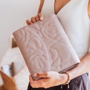 May include: A light pink clutch bag with a floral pattern. The bag is held by a person. The clutch has a textured surface and a stitched floral design. The colour is a soft, neutral pink.