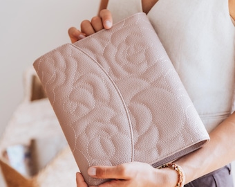 May include: A light pink clutch bag with a floral pattern. The bag is held by a person. The clutch has a textured surface and a stitched floral design. The colour is a soft, neutral pink.