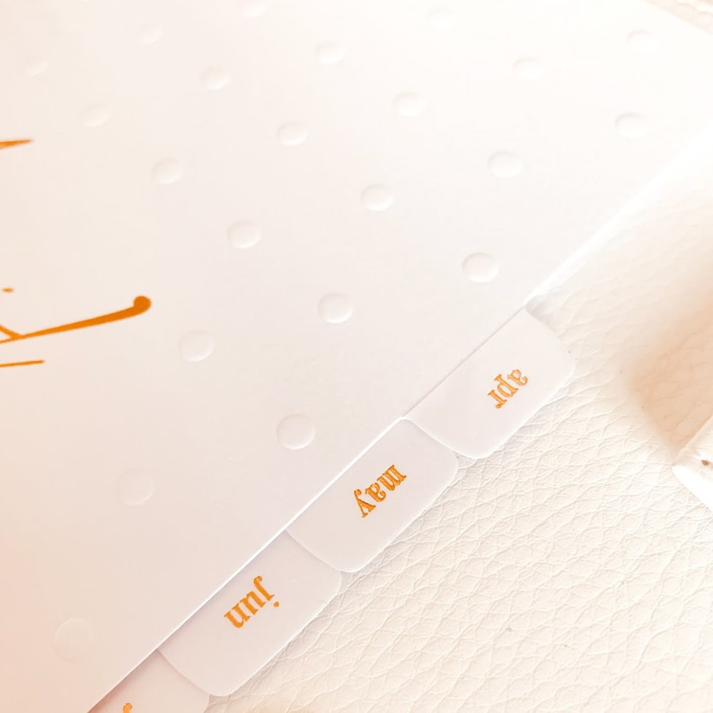 Op de afbeelding: Een witte planner met gouden tabs met de labels "Jun", "May" en "Jul".