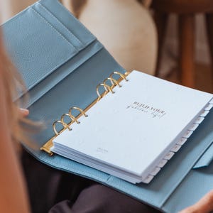 Könnte beinhalten: Ein hellblauer Ringbuchordner aus strukturiertem Leder mit goldenen Metallringen und weißen Seiten. Das Deckblatt zeigt den schwarzen Schriftzug "BUILD YOUR fabulous life!", umgeben von einem dezenten Herzmuster und Registerkarten.