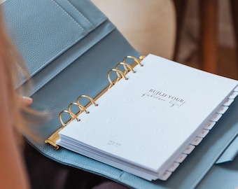 May include: A light blue textured leather binder with gold-toned metal rings holding white pages. The cover page features the text "BUILD YOUR fabulous life!" in black, surrounded by a subtle embossed heart pattern and side tab dividers.