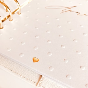 Op de afbeelding: Een witte planner met een gouden hart en gouden ringen. De planner heeft een wit stippenpatroon.