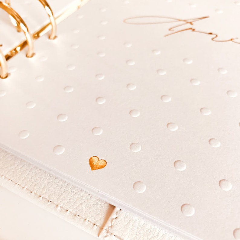 Op de afbeelding: Een witte planner met een gouden hart en gouden ringen. De planner heeft een wit stippenpatroon.