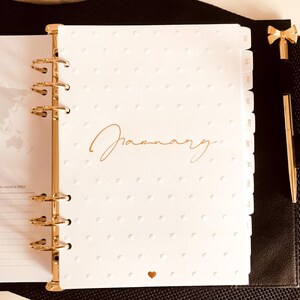 May include: A white planner with gold rings and tabs for each month of the year. The planner has a white polka dot pattern and the word "January" is written in gold script on the front cover.