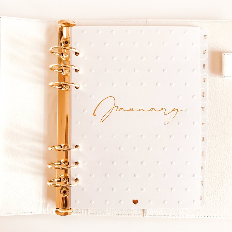 Op de afbeelding: Een witte planner met gouden ringen en een gouden inscriptie die "Januari" zegt. De planner heeft een witte achtergrond met kleine witte stippen.