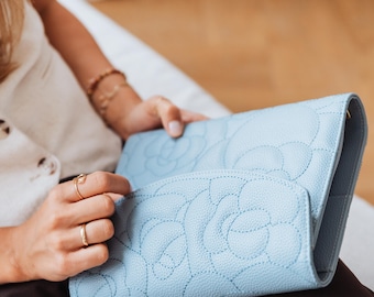 May include: A pale blue clutch bag with a floral design. The bag has a textured finish and a quilted pattern with stitched detailing. The clutch is open, showing the inside. The overall look is elegant and stylish.