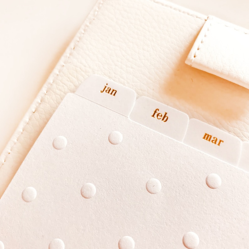 Op de afbeelding: Een witte planner met gouden tabbladen met de labels "Jan", "Feb" en "Mar".