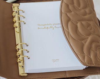 May include: A tan, quilted leather-like planner with a gold-toned six-ring binder mechanism. The white interior pages feature embossed floral patterns and the gold-printed text: Thoughtfully planned, beautifully lived. Oh So Bella my planner.