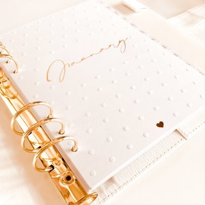 Op de afbeelding: Een witte planner met gouden ringen en een gouden stippenpatroon. Het woord "Januari" staat in gouden letters op de omslag. Een klein gouden hartje bevindt zich in de rechterbenedenhoek.