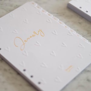 Może przedstawiać: Biała strona planera z wytłoczonym wzorem serca i napisem "January" złotym pismem. Strona ma serię zakładek i jest częścią większego planera lub notatnika. Projekt jest prosty i elegancki.