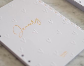 Op de afbeelding: Witte plannerpagina met een reliëf hartpatroon en het woord "January" in gouden script. De pagina heeft een reeks tabbladen en maakt deel uit van een grotere planner of notitieboek. Het ontwerp is eenvoudig en elegant.