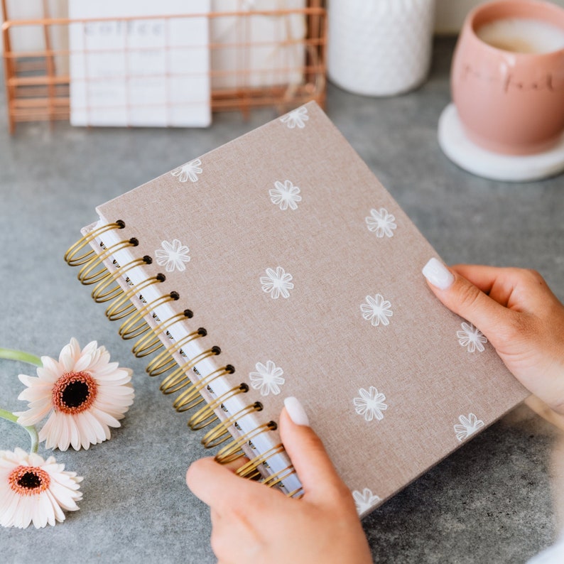 Girl is holding planner in her hands, on the side on the table is mug with coffee and pink flower. Planner have a floral designer and gold wire.