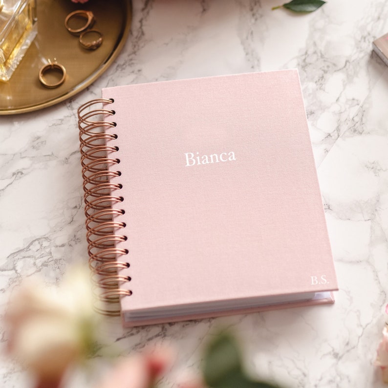 pink planner is on the white marble table, on the side gold tray with three gold rings and perfiumes in glass bottle.