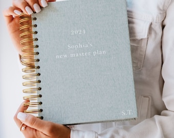 Könnte beinhalten: Ein hellblauer Planer mit einer goldenen Spiralbindung. Der Einband trägt den Text "2024 Sophia's new master plan" und "S.T." in weißer Schrift.