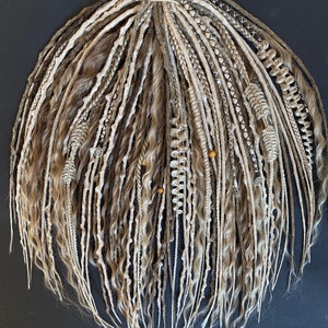 Può includere: Un set di dreadlocks fatti a mano in tonalità marroni e bionde. I dreadlocks variano nella consistenza, alcuni sono lisci e altri ondulati. Alcuni dreadlocks sono decorati con perline e ciondoli. Lo sfondo è nero.