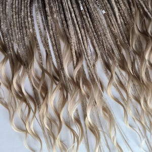 May include: Close-up of a bundle of dreadlocks with a gradient effect. The top portion is a dark brown color, transitioning to a lighter, sandy blonde at the ends. The ends of the dreadlocks have a wavy, curly texture.