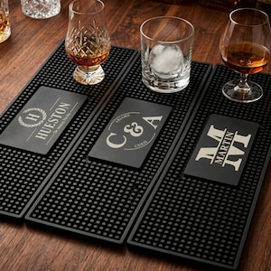 May include: Three rectangular black rubber bar mats with raised circular nubs. Each mat features a personalized center panel: one with HUESTON and a laurel wreath, one with C & A, and one with the name MARTIN in large block letters.