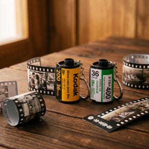 Pode incluir: Dois porta-chaves de rolo de filme de 35mm, um amarelo Kodak e um verde Fujifilm, cada um com um anel de metal. Aos rolos estão presas tiras de filme de 35mm com fotografias impressas, com tiras adicionais visíveis nas proximidades.