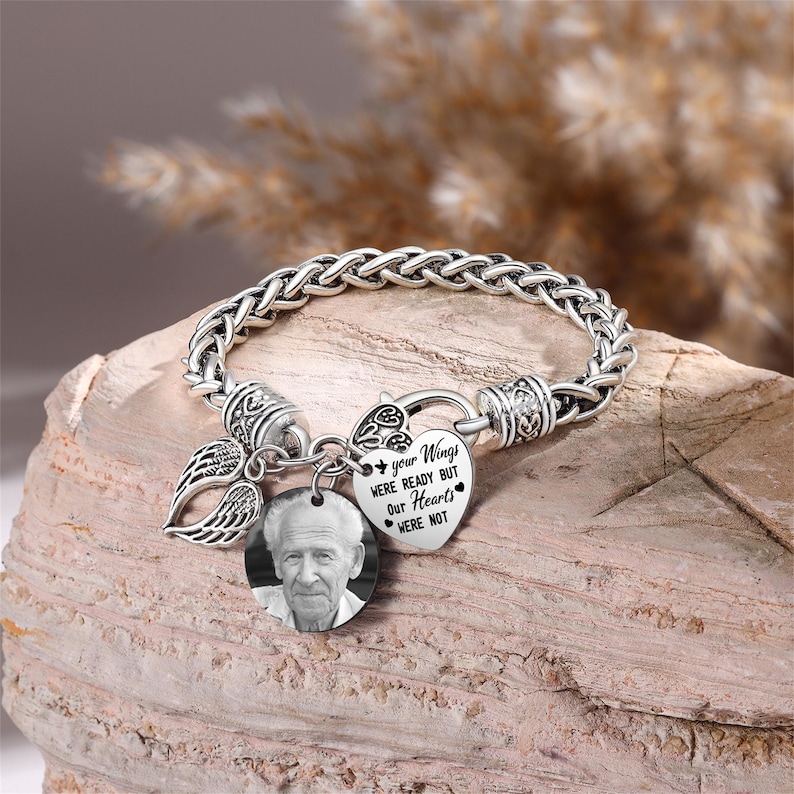 Puede incluir: Pulsera de plata con un dije en forma de coraz&oacute;n que dice "Tus alas estaban listas pero nuestros corazones no". El dije est&aacute; unido a un dije de ala de &aacute;ngel de plata y a un dije de foto redondo con una foto en blanco y negro de un hombre mayor.