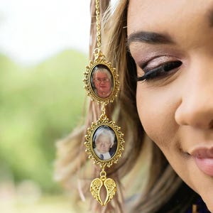 May include: A gold-toned metal charm accessory featuring two vertical oval frames with ornate scalloped edges, each containing a small portrait photograph of an individual, finished with a pair of small gold-toned angel wings at the bottom.