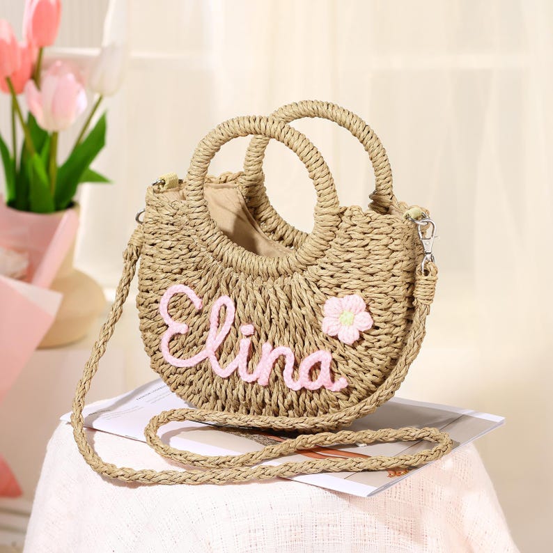 May include: A straw handbag with a pink flower and the name "Elina" embroidered on the front. The bag has a top handle and a long strap.