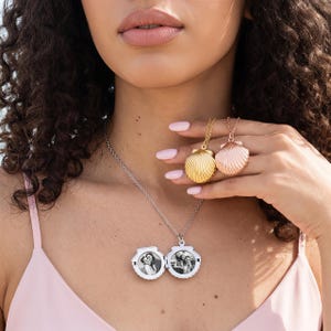 Könnte beinhalten: Eine silberfarbene muschelförmige Medaillon-Halskette mit zwei runden Schwarz-Weiß-Fotos im Inneren. Das Medaillon hängt an einer feinen Kette. Zwei weitere muschelförmige Anhänger in Gold- und Roségoldtönen werden in der Hand gehalten.