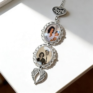 May include: A silver-toned hanging ornament featuring two oval frames with ornate filigree borders, each containing a photograph. At the top is a heart-shaped charm engraved with 2026, and at the bottom is a pair of angel wings charm.