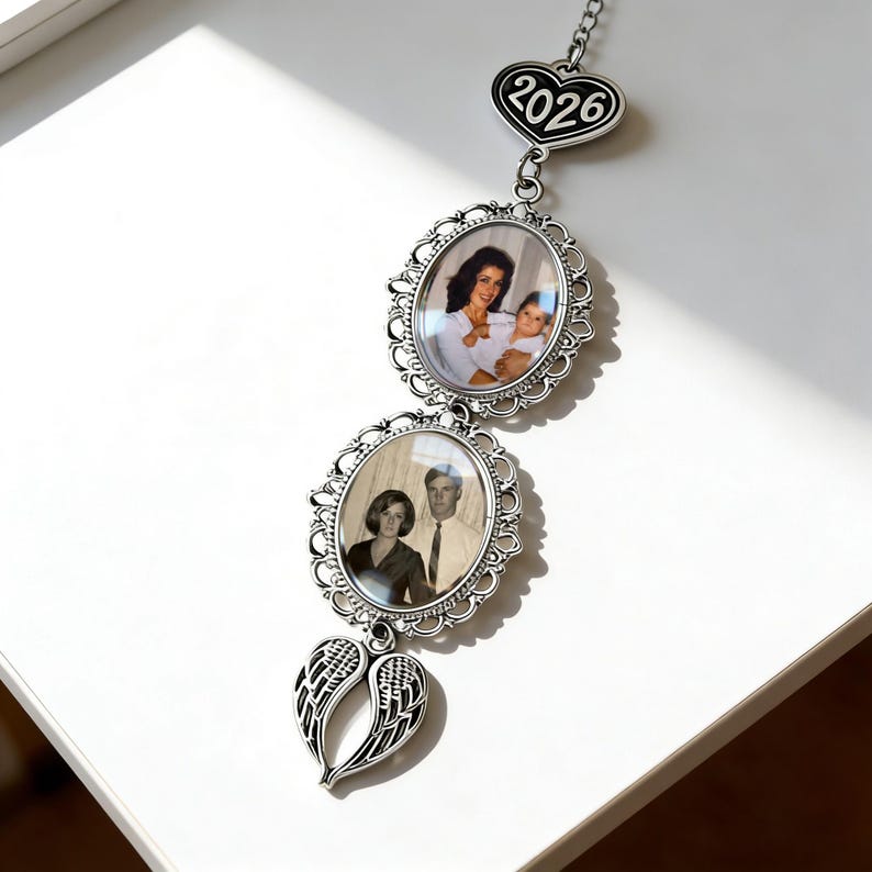 May include: A silver-toned hanging ornament featuring two oval frames with ornate filigree borders, each containing a photograph. At the top is a heart-shaped charm engraved with 2026, and at the bottom is a pair of angel wings charm.