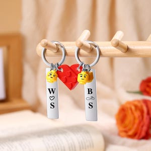 May include: Two keychains with silver rings, each with a rectangular tag and a Lego-style head charm. One tag has the letters "W", a heart, and "S". The other tag has "B", an infinity symbol, and "S". A red heart is between the tags.