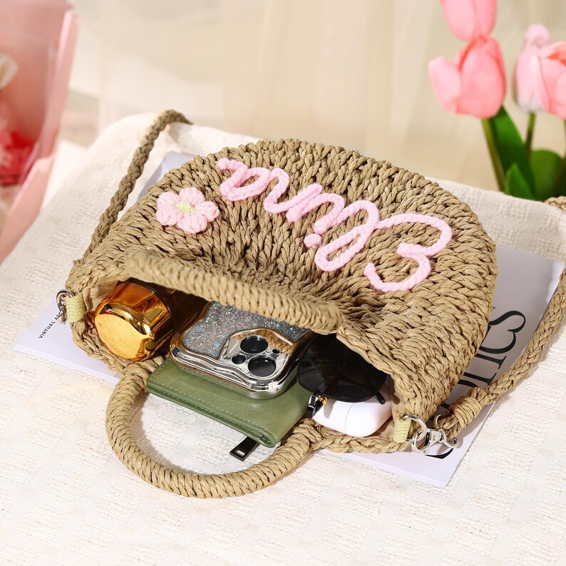 May include: A straw tote bag with a pink flower and the word "Emma" embroidered on the front. The bag is open and has a phone, sunglasses, and a small bottle inside.