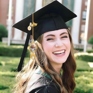 May include: A black graduation cap with a black tassel and a gold-toned metal charm attached. The charm features a small oval frame containing a photograph of a person, a circular disc, and a small gold-toned banner with the text 2024 MILES.