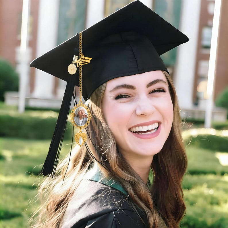 May include: A black graduation cap with a black tassel and a gold-toned metal charm attached. The charm features a small oval frame containing a photograph of a person, a circular disc, and a small gold-toned banner with the text 2024 MILES.