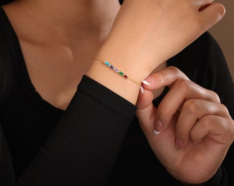 May include: A gold chain bracelet with a row of colorful square beads. The bracelet is on a person's wrist.