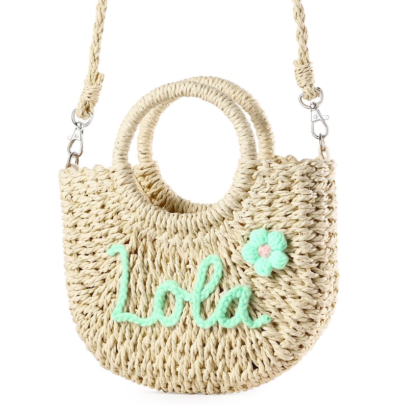 May include: A straw tote bag with a green flower and the name "Lola" embroidered on the front. The bag has a top handle and a shoulder strap.