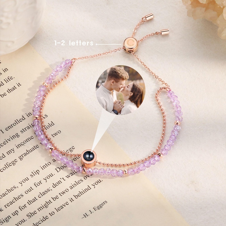 May include: A rose gold bracelet with a small, round, clear stone that projects an image. The bracelet is made of two strands of small, purple beads. The bracelet has a sliding clasp. The image projected by the stone is of a couple kissing.